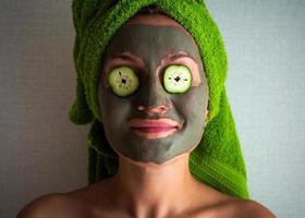 bellissimo giovane donna con facciale maschera su sua viso Tenere fette di cetriolo. foto