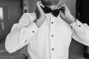 vicino su di un' uomo con bianca camicia fare un' preparazione e posa indossare un' arco cravatta nel monocromatico immagine. foto