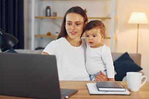 seduta di il tavolo con il computer portatile. madre con sua poco figlia è a casa insieme foto