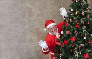 Santa Claus è festeggiare Natale nel felicità e eccitazione mentre nascondiglio dietro a completamente decorato Natale albero e agitando per stagione saluto e felicità concetto foto