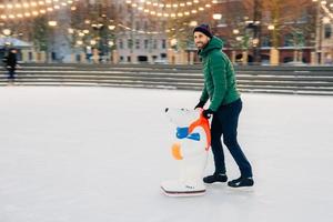 Il pattinatore maschio contento di essere sulla pista di pattinaggio, usa l'ausilio per lo skate mentre cerca di essere in equilibrio, guarda felicemente da parte, posa sul ghiaccio. uomo allegro vestito con abiti caldi, coinvolto in uno stile di vita attivo. foto