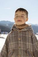 ritratto di poco adorabile ragazzo con inverno paesaggio nel il sfondo. carino, sorridente caucasico bambino con Marrone cappotto in piedi all'aperto e godendo bene tempo atmosferico, blu cielo, neve. Buona, positivo umore. foto