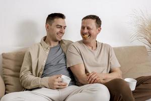 contento gay coppia con casuale Abiti la spesa tempo insieme a casa, Guardando film su il il computer portatile e potabile tè. Due caucasico uomini rilassante. omosessuale relazioni e alternativa amore. foto