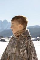 ritratto di poco adorabile ragazzo con inverno paesaggio nel il sfondo. carino, sorridente caucasico bambino con Marrone cappotto in piedi all'aperto e godendo bene tempo atmosferico, blu cielo, neve. Buona, positivo umore. foto