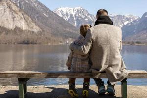 padre e figlio seduta insieme all'aperto su il panchina e godendo montagne, neve, bene tempo atmosferico, blu cielo. poco ragazzo e il suo padre la spesa tempo insieme. famiglia volta. bellissimo inverno paesaggio. foto