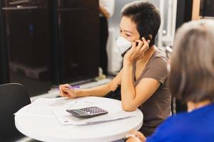 donna indossare viso maschera parlando su cellula Telefono Tenere penna con lavoro d'ufficio su tavoletta. foto