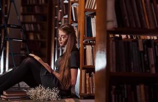 si siede su il pavimento. femmina alunno è nel biblioteca quello pieno di libri. concezione di formazione scolastica foto