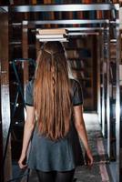 allegro ragazza avere divertimento mentre mette libri su sua testa. femmina alunno è nel biblioteca. concezione di formazione scolastica foto