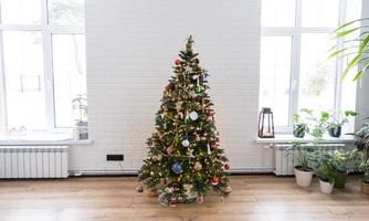 Natale albero nel il bianca interno di un' Casa con grande finestre. raggiante Fata luci ghirlande interno decorazione di il studio camera. in vaso impianti nel il casa foto