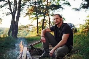 tempo per un' riposo. uomo nel nero camicia vicino il fuoco di bivacco nel il foresta a il suo fine settimana tempo foto