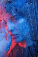 giorno di pioggia. girato in studio in uno studio buio con luce al neon. ritratto di bella ragazza dietro il vetro bagnato foto