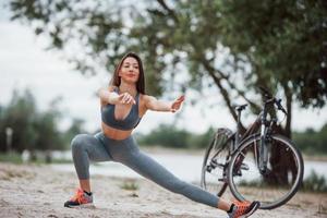 fare squat. ciclista femminile con una buona forma del corpo che fa esercizi di yoga e si allunga vicino alla sua bici sulla spiaggia durante il giorno foto