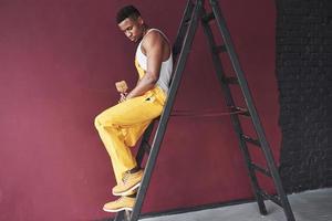 si siede su scala a pioli. giovane africano americano lavoratore nel il giallo uniforme avere alcuni lavoro foto