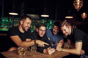 divertente soddisfare su smartphone. tre amici riposo nel il pub con birra nel mani. avendo conversazione foto