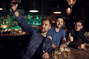 fabbricazione autoscatto. tre gli sport fan nel un' bar Guardando calcio. con birra nel mani foto