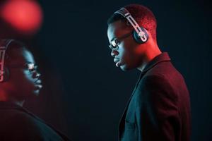 godendo ascoltando musica nel cuffia. nel occhiali. futuristico neon illuminazione. giovane africano americano uomo nel il studio foto