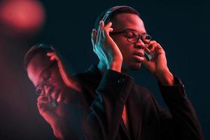 godendo ascoltando musica nel cuffia. nel occhiali. futuristico neon illuminazione. giovane africano americano uomo nel il studio foto