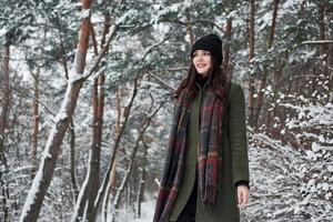 in piedi nel il mezzo di il boschi. allegro giovane ragazza nel caldo Abiti avere un' camminare nel il inverno foresta a giorno foto