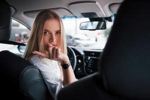 trasformato indietro per il cameraman a sedile posteriore. bellissimo bionda ragazza seduta nel il nuovo auto con moderno nero interno foto