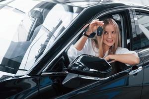 soddisfatto proprietario. bellissimo bionda ragazza seduta nel il nuovo auto con moderno nero interno foto