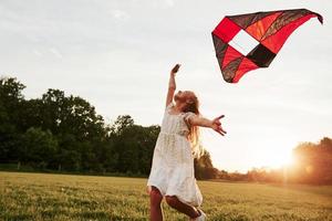 sensazione libertà. contento ragazza nel bianca Abiti avere divertimento con aquilone nel il campo. bellissimo natura foto