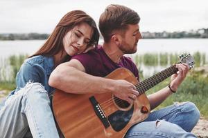 sensazione Buona. uomo giochi chitarra per il suo fidanzata a spiaggia su loro picnic a giorno foto