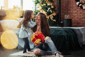 grato ragazza è dando un' bacio. madre e figlia si siede nel vacanza decorato camera e detiene regalo scatola foto