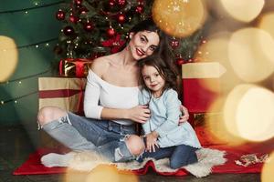 guardare in il telecamera. allegro madre e figlia seduta vicino il Natale albero quello dietro. carino ritratto foto