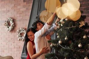 ragazza abbracciare sua fidanzato con amore. romantico coppia condimento su Natale albero nel il camera con Marrone parete e camino foto