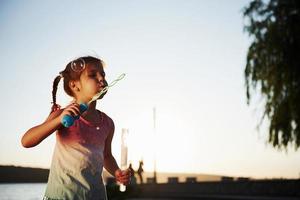 sera volta. contento poco ragazza giocando con bolle vicino il lago a parco foto