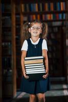 carino poco ragazza nel bicchieri sta nel il biblioteca pieno di libri. concezione di formazione scolastica foto