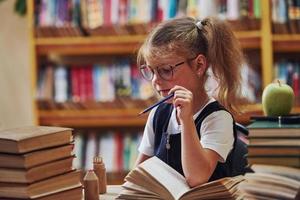 carino poco ragazza con trecce è nel il biblioteca. Mela su il libri foto