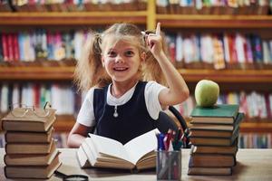 io avere un idea. carino poco ragazza con trecce è nel il biblioteca. Mela su il libri foto