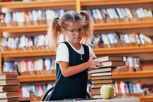 carino poco ragazza con trecce è nel il biblioteca. Mela su il libri foto