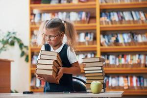 carino poco ragazza con trecce è nel il biblioteca. Mela su il libri foto