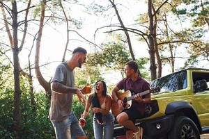 cantando canzoni insieme. amici avere simpatico fine settimana all'aperto vicino il loro verde auto con acustico chitarra foto