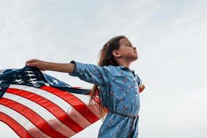 patriottico femmina ragazzo con americano bandiera nel mani. contro nuvoloso cielo foto