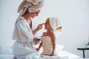 utilizzando crema per chiaro pelle. giovane madre con sua figlia avere bellezza giorno in casa nel bianca camera foto