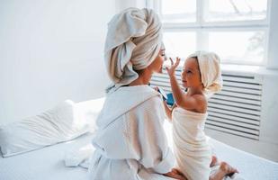 utilizzando crema per chiaro pelle. giovane madre con sua figlia avere bellezza giorno in casa nel bianca camera foto