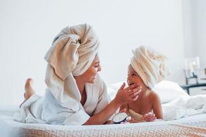 utilizzando crema. giovane madre con sua figlia avere bellezza giorno in casa nel bianca camera foto