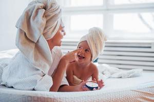 utilizzando crema. giovane madre con sua figlia avere bellezza giorno in casa nel bianca camera foto