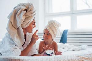 utilizzando crema. giovane madre con sua figlia avere bellezza giorno in casa nel bianca camera foto