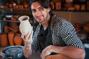 giovane ceramista detiene fresco fatto a mano pentola nel mano. soddisfatto con risultati di il suo opera foto