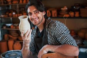 giovane ceramista detiene fresco fatto a mano pentola nel mano. soddisfatto con risultati di il suo opera foto