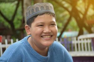 ritratto asiatico musulmano o islamico ragazzo seduta nel il scuola parco e sorridente felicemente, morbido e selettivo messa a fuoco. foto