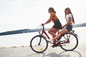 Due femmina amici su il bicicletta avere divertimento a spiaggia vicino il lago foto