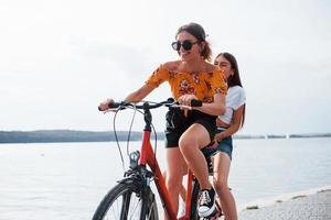 Due femmina amici su il bicicletta avere divertimento a spiaggia vicino il lago foto