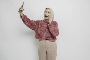 un' ritratto di un' contento asiatico donna indossare un' rosa maglione e hijab, Tenere sua Telefono, isolato di bianca sfondo foto