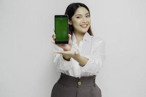 eccitato asiatico donna indossare bianca camicia puntamento a il copia spazio accanto sua mentre Tenere sua Telefono, isolato di bianca sfondo foto