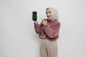 eccitato asiatico musulmano donna indossare rosa maglione e hijab puntamento a il copia spazio accanto sua mentre Tenere sua Telefono, isolato di bianca sfondo foto